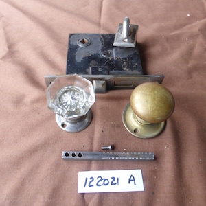 May include: A vintage door hardware set with a brass knob, a glass crystal knob, a black metal door plate, and a metal latch. The set also includes a metal rod and a screw.