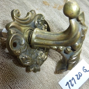 Może przedstawiać: Ozdobny mosiężny klamka do drzwi z dekoracyjnym wzorem kwiatowym i haczykowatym uchwytem. Klamka jest przymocowana do metalowej płytki za pomocą śruby. Klamka jest oznaczona "727 20 G".