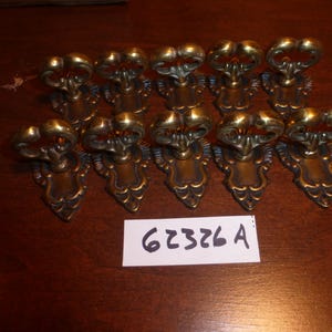 Könnte beinhalten: Ein Set aus zehn Messing-Schubladengriffen mit verzierten, geschwungenen Griffen und dekorativen Rückplatten. Jeder Griff hat eine polierte Metalloberfläche. Ein weißes rechteckiges Etikett mit dem handgeschriebenen Text 62326 A liegt darunter.