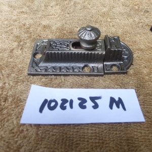 May include: An antique metal door latch with an ornate design. The latch has a decorative knob and a rectangular base with intricate patterns. The text "102125 M" is on a white paper below the latch.