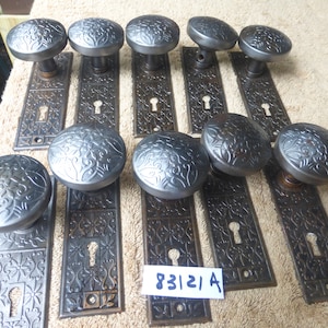 Op de afbeelding: Tien antieke deurknoppen met sierlijke bloemmotieven. De knoppen zijn van metaal en hebben een donkere, verweerde afwerking. De achterplaten zijn ook van metaal en hebben een vergelijkbaar ontwerp. De knoppen zijn gerangschikt in twee rijen van vijf. De tekst "83121 A" is zichtbaar in de afbeelding.