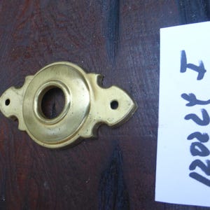 May include: A gold-toned metal door knob cover with a decorative design. The cover has a round opening in the centre and two small holes on either side. The cover is on a dark brown wooden surface.