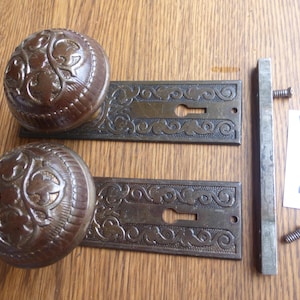 May include: Two round, dark brown door knobs with embossed floral patterns, each attached to a rectangular metal backplate featuring intricate scrollwork and a keyhole. Includes a square metal spindle, two screws, and a white tag labeled 30324 E.