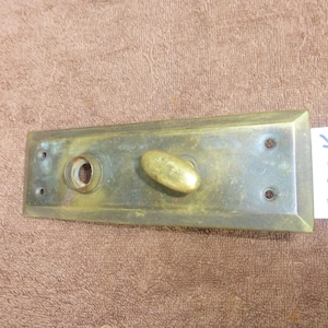 May include: Antique brass door hardware, featuring a rectangular backplate with a keyhole and an oval-shaped handle. The hardware has a weathered appearance, with visible signs of age and use. A small white tag with the text "92823 K" is attached.
