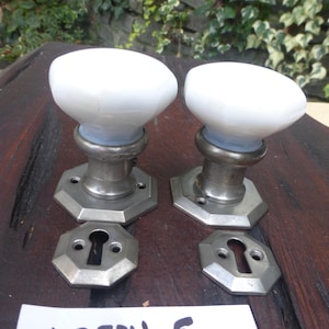 Könnte beinhalten: Zwei antike weiße Porzellanknopf-Türgriffe mit silbernen Metallsockeln und Schlüssellöchern. Die Platten sind achteckig. Der Text "112524 E" ist auf einem weißen Etikett geschrieben.