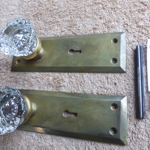 May include: Two antique brass door knobs with clear glass crystal knobs. The knobs are attached to rectangular plates with keyholes. The plates have two screws each. A piece of paper with the text "10524B" is visible in the image.