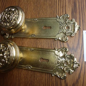 May include: Two matching brass door knobs with ornate, circular handles and rectangular backplates featuring intricate scrollwork and a central keyhole. A small white paper tag with the handwritten text 30224 J is placed to the right.