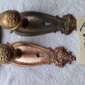 May include: Two antique door handle sets with ornate floral designs. The handles are oval-shaped and made of brass, with a key and a keyhole. The backplates are a mix of bronze and copper tones. A tag with the number "1219" is visible.