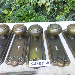 May include: Five antique brass door knobs with matching backplates. The knobs are round with a decorative top, and the backplates have keyholes and ornate detailing. The finish varies from dark to a lighter brass colour. A tag with the text "52125 M" is visible.