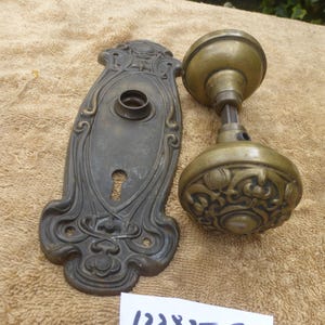 May include: Antique brass door hardware set, including a decorative backplate with a keyhole and a detailed doorknob. The doorknob features an ornate floral design. The set is aged with a patina and includes the number "122825 G".