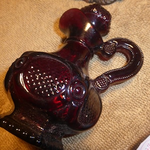 Puede incluir: Una jarra de vidrio rojo oscuro con un asa decorativa y una superficie texturizada. La jarra tiene un pequeño pico y una parte superior redonda.