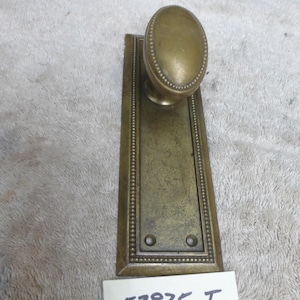 May include: Antique brass door knob with an oval handle and rectangular backplate. The backplate and handle are adorned with a beaded border. The text "52825 I" is printed on a white label.