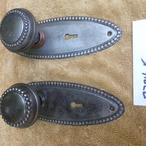 May include: Two antique door knobs with oval escutcheons. The knobs and escutcheons are made of metal and have a dark gray finish. The escutcheons have a beaded edge and a keyhole. The knobs have a beaded design around the edge.