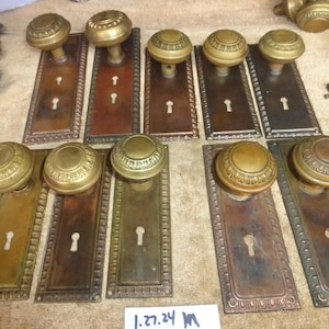 Op de afbeelding: Een verzameling antieke messing deurknoppen en platen. De knoppen zijn rond met decoratieve details en de platen zijn rechthoekig met sleutelgatuitsparingen. Het metaal heeft een warme, verouderde patina met verschillende tinten bruin en goud.