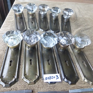 Könnte beinhalten: Zehn antike Messing-Türknäufe mit durchsichtigen Kristallknäufen aus Glas. Die Knäufe sind in einer Reihe auf einer braunen Stoffoberfläche angeordnet. Die Knäufe haben ein Schlüsselloch und sind in gutem Zustand. Der Text "122820 B" ist auf einem weißen Aufkleber geschrieben.