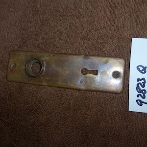 Może przedstawiać: Prostokątna mosiężna płyta drzwiowa z otworem na klucz i okrągłym otworem. Płyta ma zużyty, postarzany wygląd. Biała etykieta z napisem "92823 Q" znajduje się obok płyty.