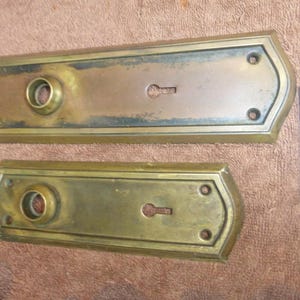 May include: Two antique brass door plates. The larger plate has a keyhole and a circular opening. The smaller plate has a keyhole and a circular opening. Both plates have a rectangular shape with rounded edges and screw holes. A white tag with the number "92823 Z" is on the right.