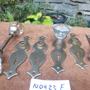 May include: Set of antique door knobs and backplates. The knobs are made of clear glass and the backplates are brass. The backplates have a keyhole and a decorative design. The set includes the text "100423 F".