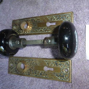 May include: Antique brass door hardware set with two ornate plates and two black ceramic door knobs. The plates have a floral design and the knobs have a smooth finish. The set is labeled "8132/B".
