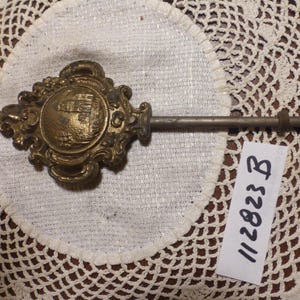 May include: An antique brass clock key with an ornate, decorative face featuring a circular emblem. The key has a long, slender shaft and is set against a white doily. A tag with the number "112823 B" is visible.