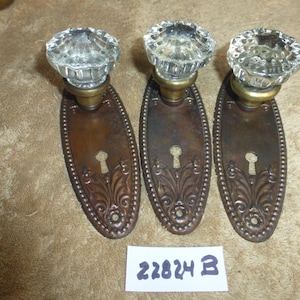 May include: Three antique door knobs with clear faceted glass knobs and ornate bronze backplates. The backplates have a keyhole and decorative floral design. The number "22824 B" is printed on a white label.