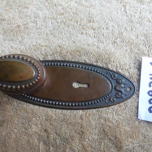May include: An antique bronze door knob and backplate. The oval knob is surrounded by small, raised dots. The backplate has a keyhole and decorative scrollwork. The number "22824 A" is written on a white paper tag.