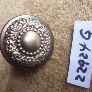 May include: A bronze-colored, circular cabinet knob with a floral and beaded design. The knob has a central, raised, circular element. A white tag with the number "22924 G" is next to the knob.