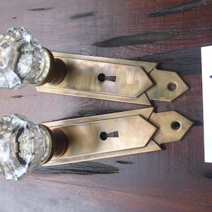 Könnte beinhalten: Zwei antike Türknauf aus Messing mit klaren Kristallknöpfen aus Glas. Die Knäufe haben ein Schlüsselloch und ein dekoratives Design. Die Knäufe befinden sich auf einer Holzoberfläche. Der Text "A 22 III III" ist auf einer weißen Karte geschrieben.