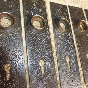 Op de afbeelding: Een verzameling antieke bronzen deurplaten. Elke plaat heeft een sierlijk, reliëf bloemenontwerp met een sleutelgat en een cirkelvormige opening voor de deurknop. Het brons heeft een donker, verouderd patina.
