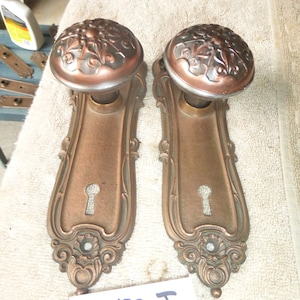 Puede incluir: Dos pomos de puerta antiguos con diseños florales ornamentados y agujeros de llave. Los pomos son de metal bronce oscuro y tienen un acabado desgastado. Los pomos están montados en placas rectangulares con bordes decorativos. Las placas también son de metal bronce y tienen un acabado desgastado. Los pomos están etiquetados con el número 81420 J.