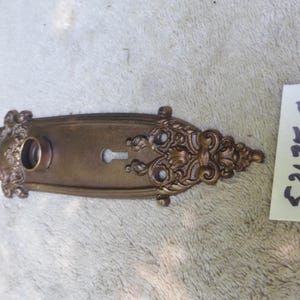 May include: Ornate antique brass door plate with keyhole and decorative floral and scrollwork. The plate has a rectangular shape with rounded ends and a circular opening. The text "52125 C" is written on a white tag.