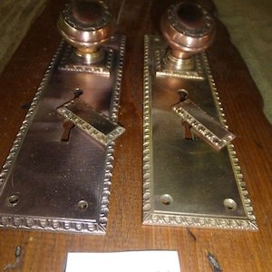 Puede incluir: Un par de pomos de puerta antiguos con placas de latón ornamentadas y detalles de cobre. Las placas tienen un borde decorativo y una cerradura. Los pomos son redondos y tienen un acabado de cobre. El texto "5421 U" está escrito en un trozo de papel blanco en la esquina inferior derecha de la imagen.