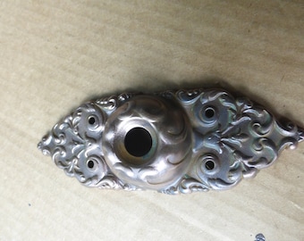 May include: Ornate, antique-style metal fixture with a central circular opening and decorative scrollwork. The fixture has a tarnished, aged appearance with a mix of silver and bronze tones, and several small holes.