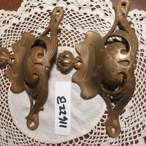 Op de afbeelding: Twee sierlijke, goudkleurige metalen decoratieve beugels met een bloemmotief. De beugels hebben een gebogen vorm en een kleine, ronde knop aan de bovenkant. De tekst "111223" is geschreven op een wit papiertje.