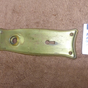 Könnte beinhalten: Eine antike Türplatte aus Messing mit Schlüsselloch und kreisförmiger Öffnung. Die Platte hat eine rechteckige Form mit abgerundeten Kanten und kleinen Schraubenlöchern. Der Text "72823 J" befindet sich auf einem weißen Etikett.