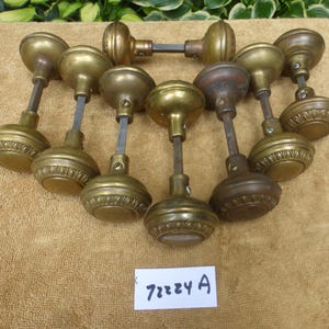 May include: A collection of eight antique brass door knobs with round, fluted bases and central spindles. Each knob features a decorative circular rim pattern. A small white rectangular tag with the handwritten black text 72224 A is included.