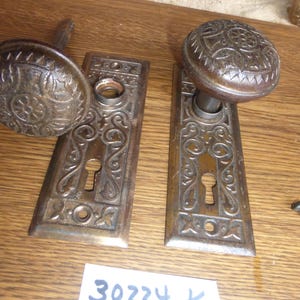May include: Two antique metal door knobs with matching rectangular backplates featuring intricate embossed scrollwork and floral patterns. Each plate includes a keyhole and screw holes. A white paper label displays the handwritten text 30224 K.