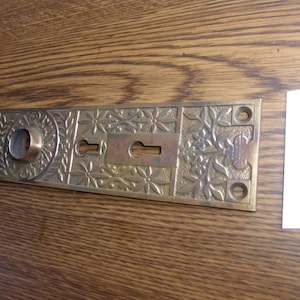 May include: An antique brass door plate with intricate floral and leaf designs. It has a keyhole and a circular opening. The rectangular plate is set on a wooden surface. The plate's dimensions are approximately 20 cm.