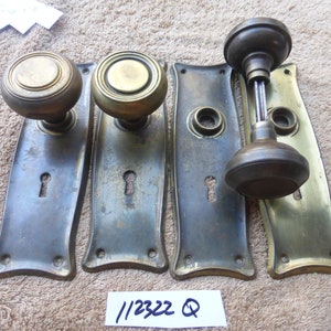 May include: Set of four antique brass door knobs and backplates. The knobs are round with a decorative rim and the backplates are rectangular with a keyhole. The backplates have a curved shape at the bottom.