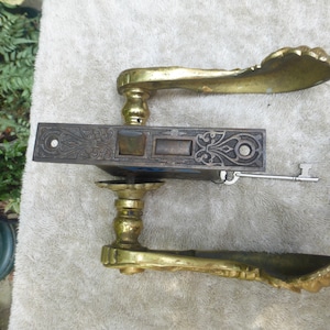 Könnte beinhalten: Antiker Türgriff-Satz aus Messing mit verzierten Details und zwei Schlüsseln. Der Griff hat ein dekoratives, geschwungenes Design. Das Schloss ist aus dunklem Metall mit aufwendigen Schnitzereien. Die Schlüssel sind silberfarben und haben ein schlichtes Design.