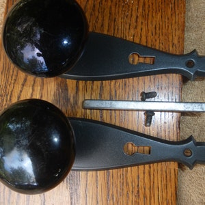 Puede incluir: Dos pomos de puerta negros con placas traseras a juego y una barra de metal, junto con dos tornillos. Los pomos son redondos y brillantes, y las placas traseras tienen un diseño de ojo de cerradura. Sobre una superficie de madera.