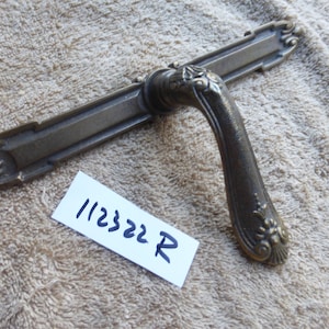Puede incluir: Una manilla de puerta vintage, adornada, de latón con una placa posterior rectangular larga. La manilla tiene un diseño floral decorativo y la placa posterior tiene un diseño geométrico simple. La manilla está etiquetada con "112322 R".