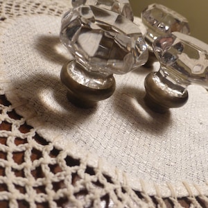 Op de afbeelding: Vier heldere glazen kastknoppen met messing bases. De knoppen hebben een facetgeslepen ontwerp en zijn gerangschikt op een wit gehaakt doily.
