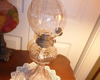 May include: A clear glass oil lamp with a swan design etched on the globe. The lamp has a decorative base and a metal burner. It sits on a brown table with a white lace doily. The lamp is a vintage item.