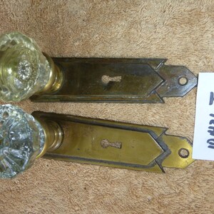 May include: Two antique brass door knobs with clear glass crystal knobs. The knobs have a keyhole and a decorative design. The knobs are on a brown surface.