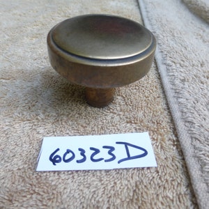 Op de afbeelding: Een ronde, messing deurknop met een verhoogde rand rond de bovenkant. De knop bevindt zich op een wit, pluche oppervlak met een wit papiertje waarop het nummer "60323D" is geschreven.