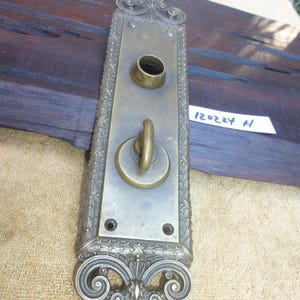 May include: Antique brass door plate with ornate details and a keyhole. The plate has a decorative design with a floral motif. The plate is mounted on a wooden surface. The text "H 12024" is written on a white label.