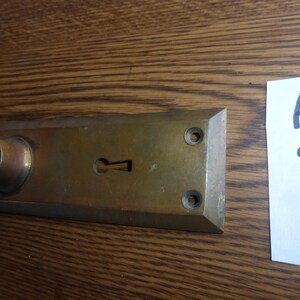 May include: A rectangular brass door plate with a keyhole and a cylindrical knob. The plate has two screw holes and is slightly tarnished. A white label with the number "121823" is next to the plate.