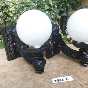 Pode incluir: Um par de arandelas de parede com bases de metal preto ornamentadas com volutas intrincadas e globos de vidro branco esféricos. Cada luminária possui uma placa traseira quadrada e um braço curvo que sustenta a cúpula redonda. Uma etiqueta diz 7826 I.