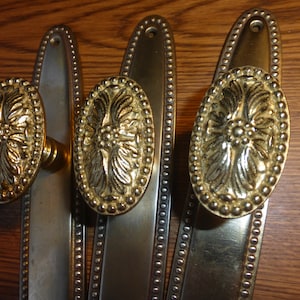 May include: Three antique brass door handles with ornate floral designs and beaded trim. The handles are oval shaped and have a polished finish.